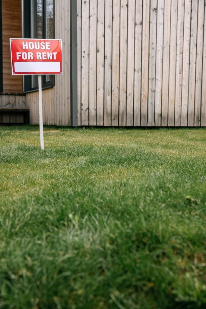 Red 'House for Rent' sign on green lawn with wooden house wall in background.